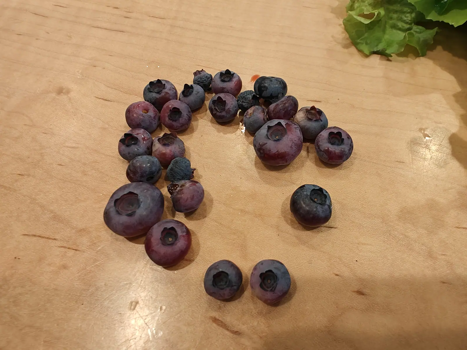 About two dozen blueberries scattered across a pale wooden board, varying in size, most still carrying a dusty grey bloom over deep purple skin.