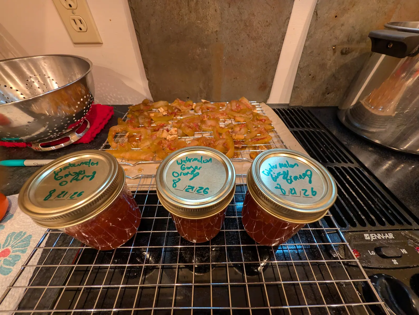 Three small jars of amber preserves with gold lids, hand-labelled in green marker, cooling on a wire rack beside a tray of dried orange-coloured pieces.