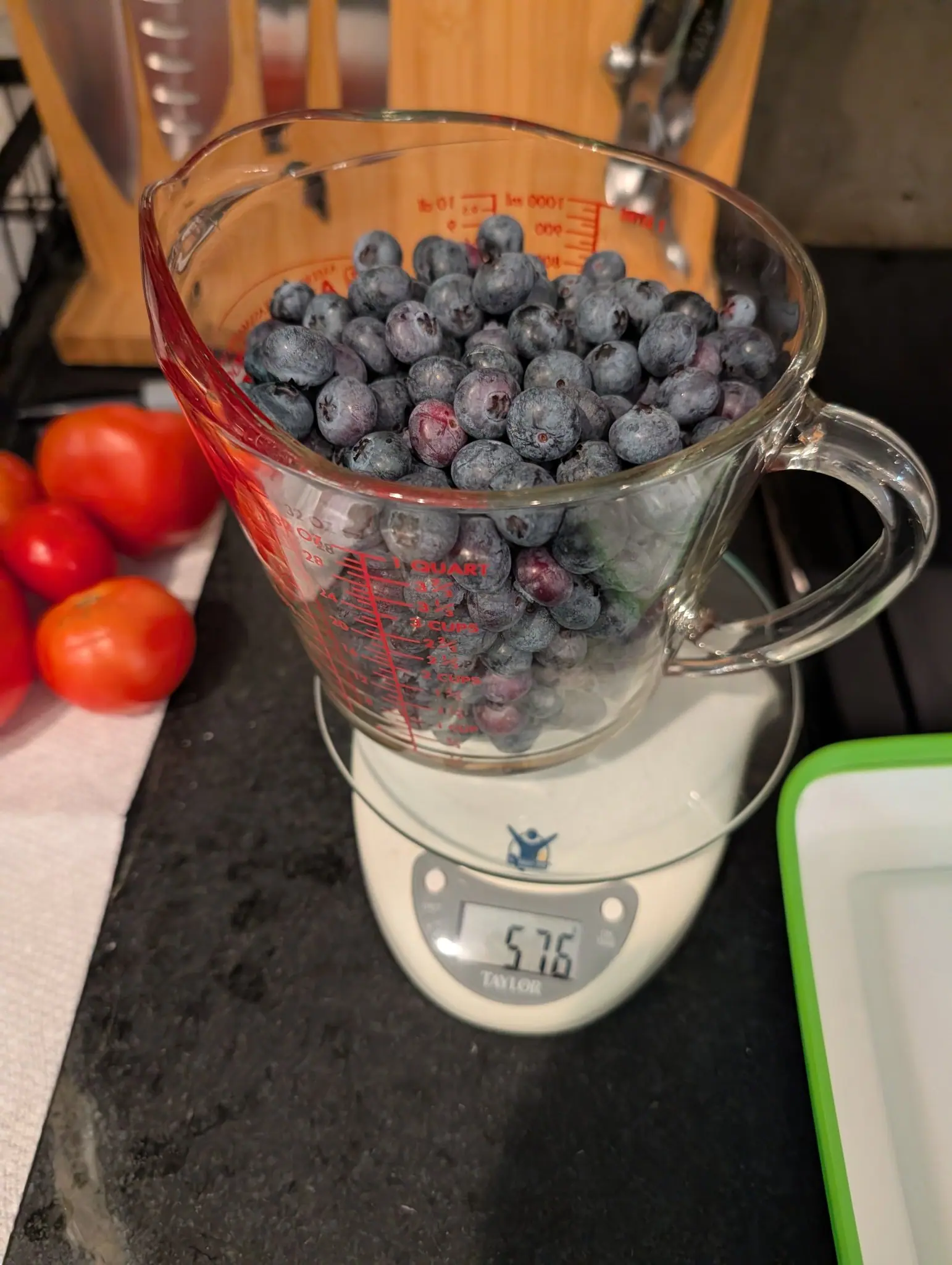 A glass measuring jug heaped with blueberries standing on a kitchen scale, its display reading 57.6. Three red tomatoes rest on a paper towel beside it.