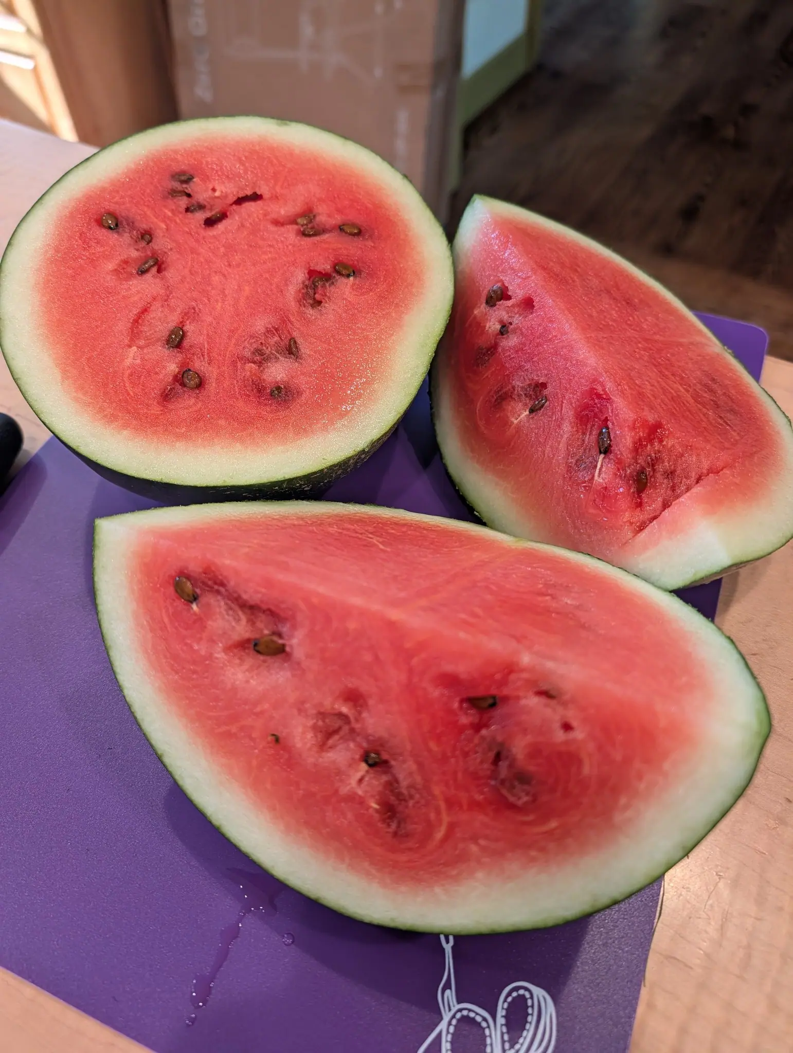 A small watermelon cut open on a purple board — one half face up and two wedges beside it. The flesh is deep pink-red and scattered with flat dark seeds, the rind pale green under a thin white layer.