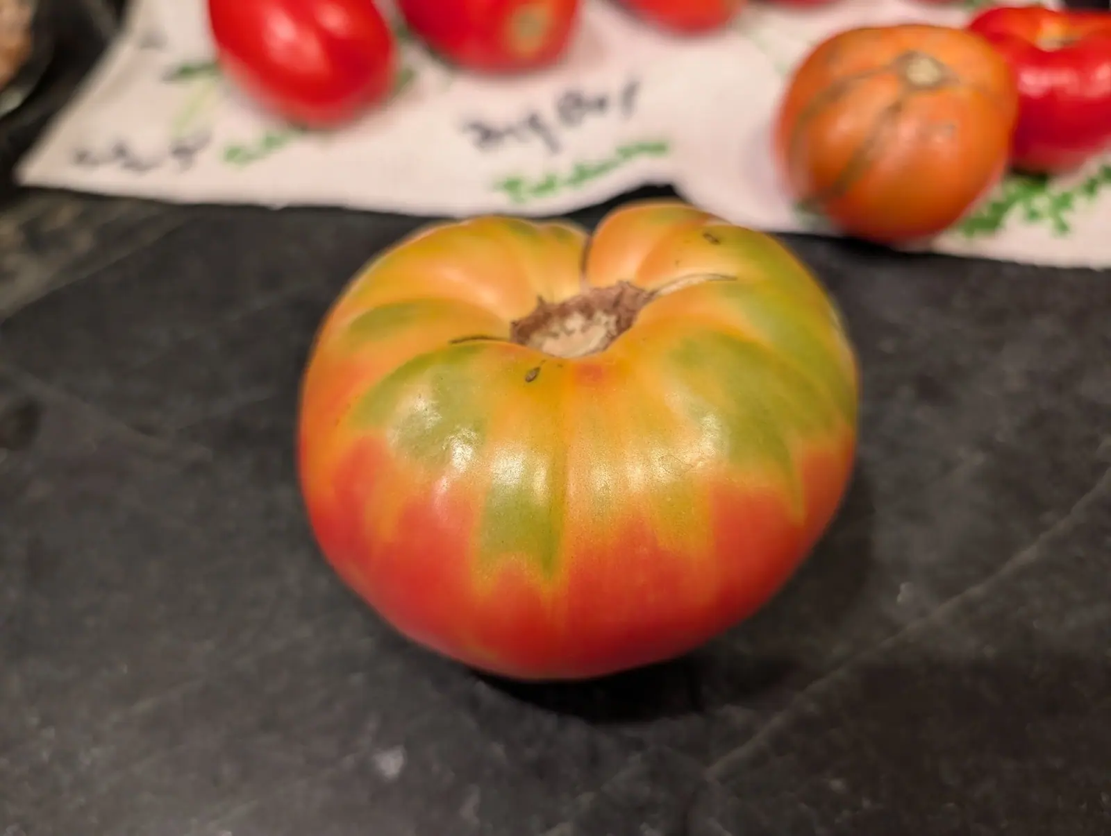 A large ribbed tomato on a dark stone worktop, its shoulders still green and shading down to orange-red around a dry stem scar, with more red fruit on a printed cloth behind.