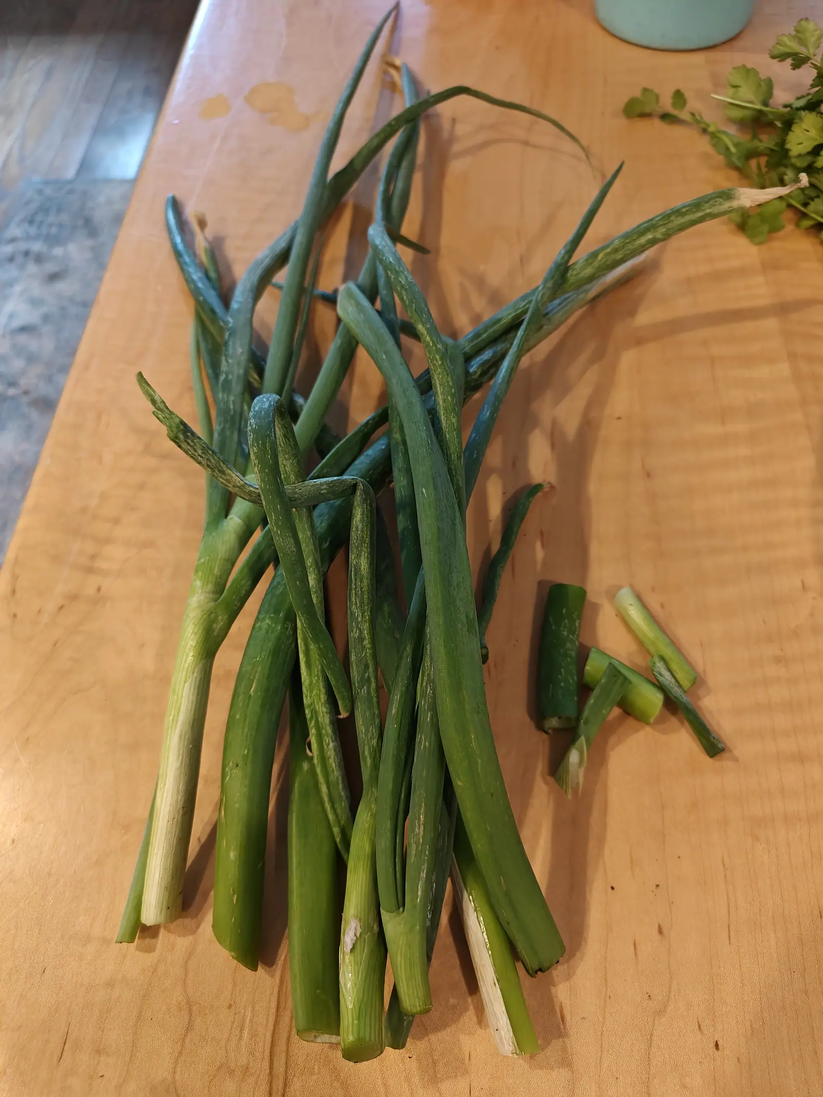 A pile of freshly pulled scallions on a pale wooden table, long green tops folding over white stems, with a few trimmed offcuts beside them and cilantro leaves at the edge.