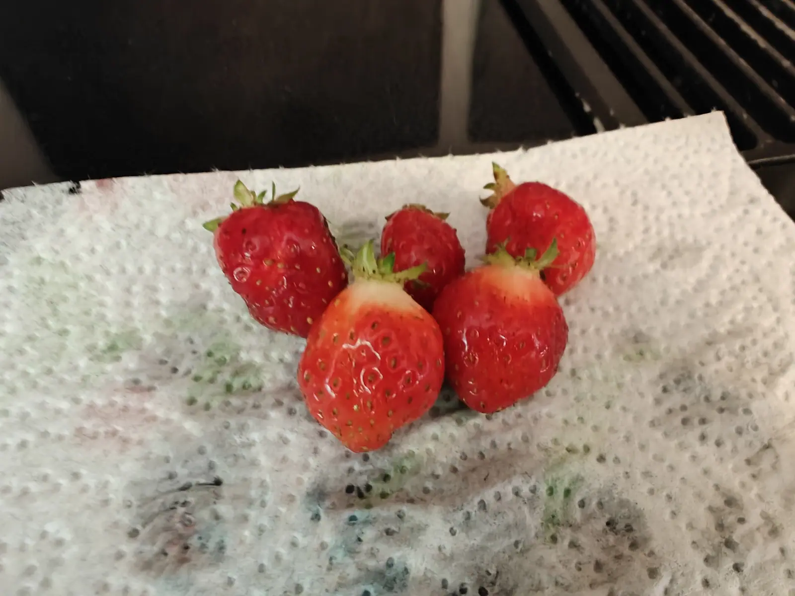 Five ripe red strawberries with their green calyces still on, grouped together on a stained paper towel set on a dark stove top.