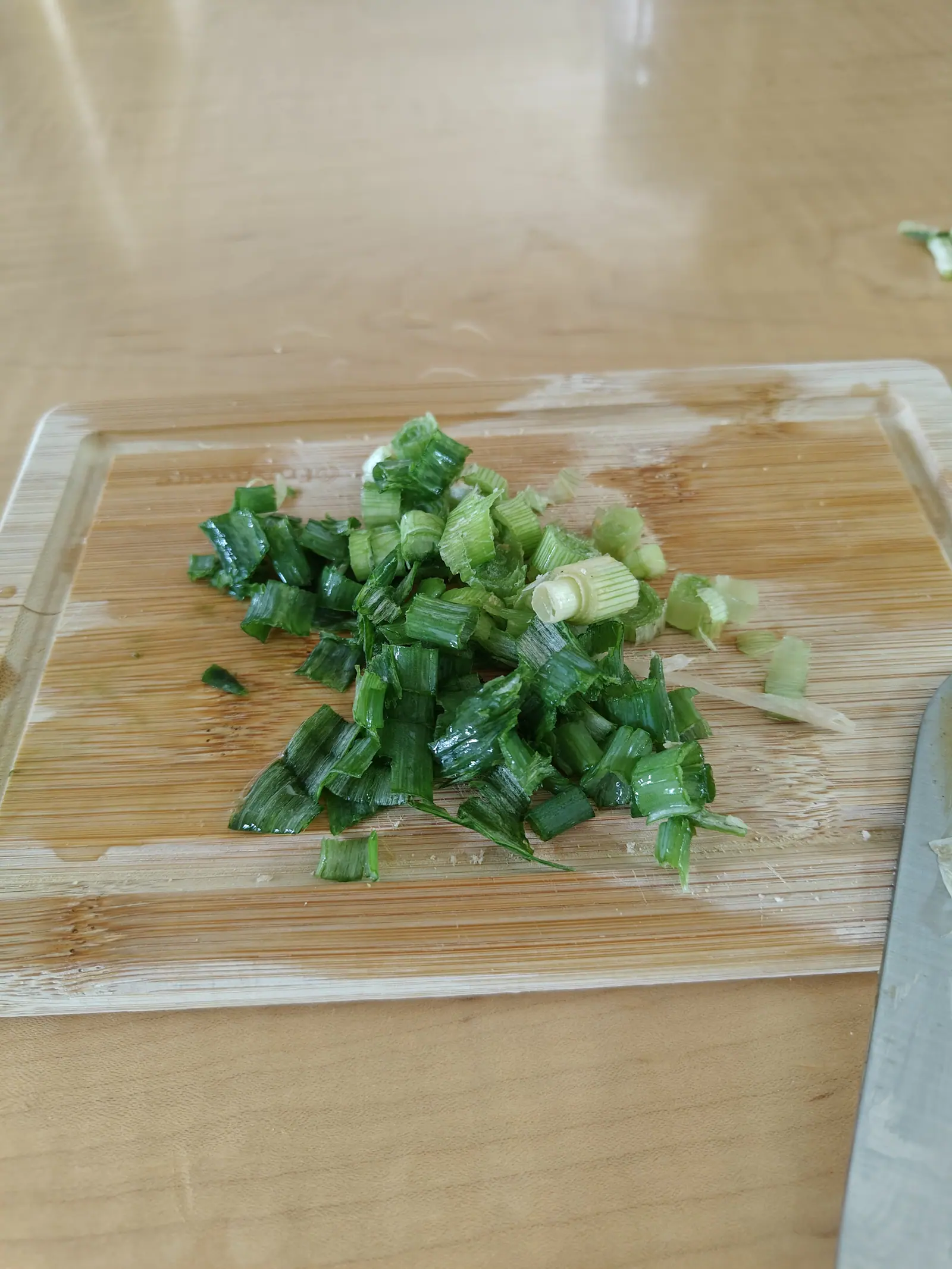 Chopped scallions heaped on a bamboo cutting board on a pale wooden table, the green rings glossy and wet, a knife blade resting at the right edge.