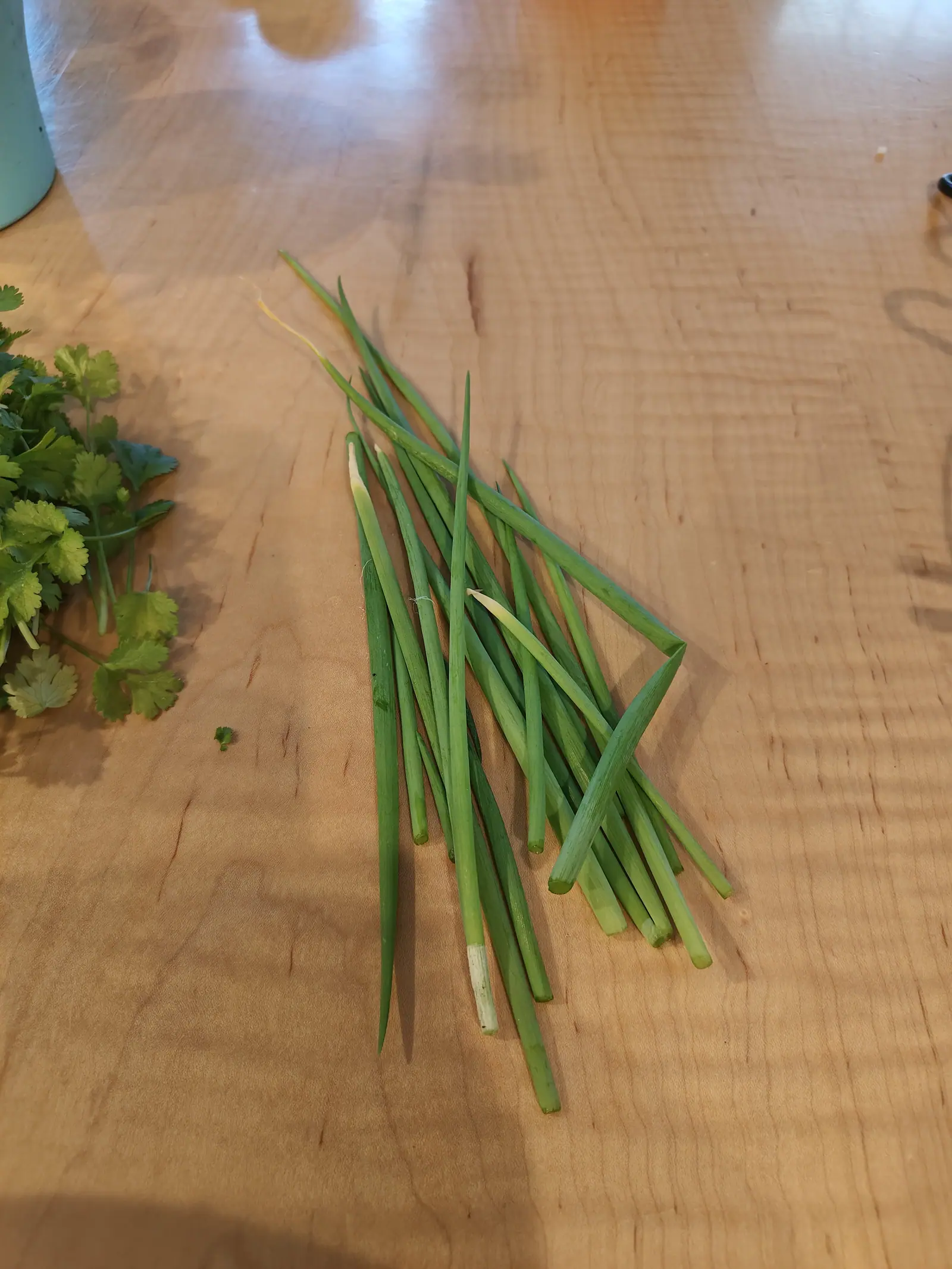 A handful of scallions laid on a pale wooden table with their white bases and green tops crossing, a loose bunch of soft green herb leaves at the left edge.