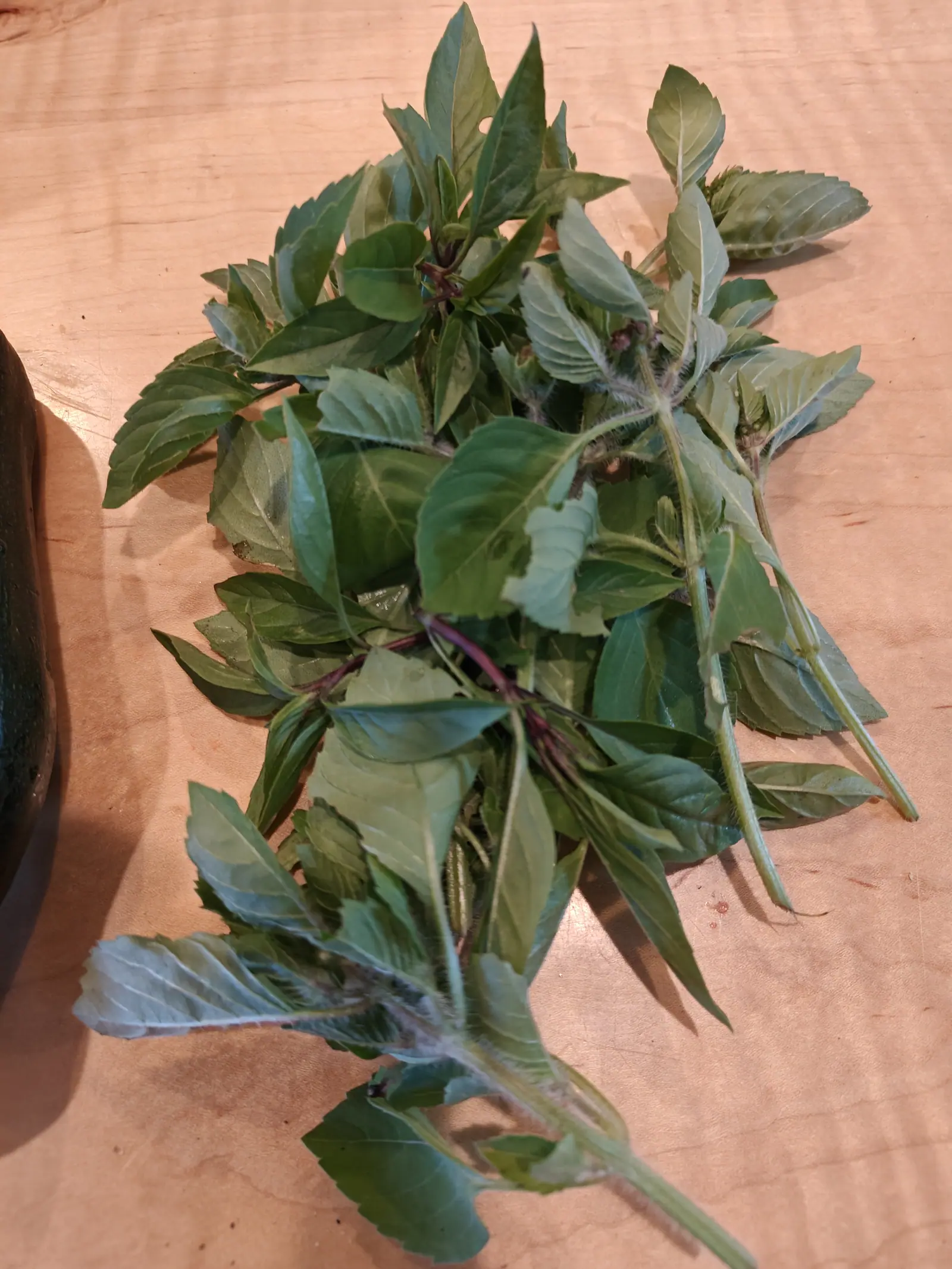 A loose handful of cut basil sprigs on a pale wooden board, green leaves on purple-flushed stems, a dark green zucchini just visible at the left edge.
