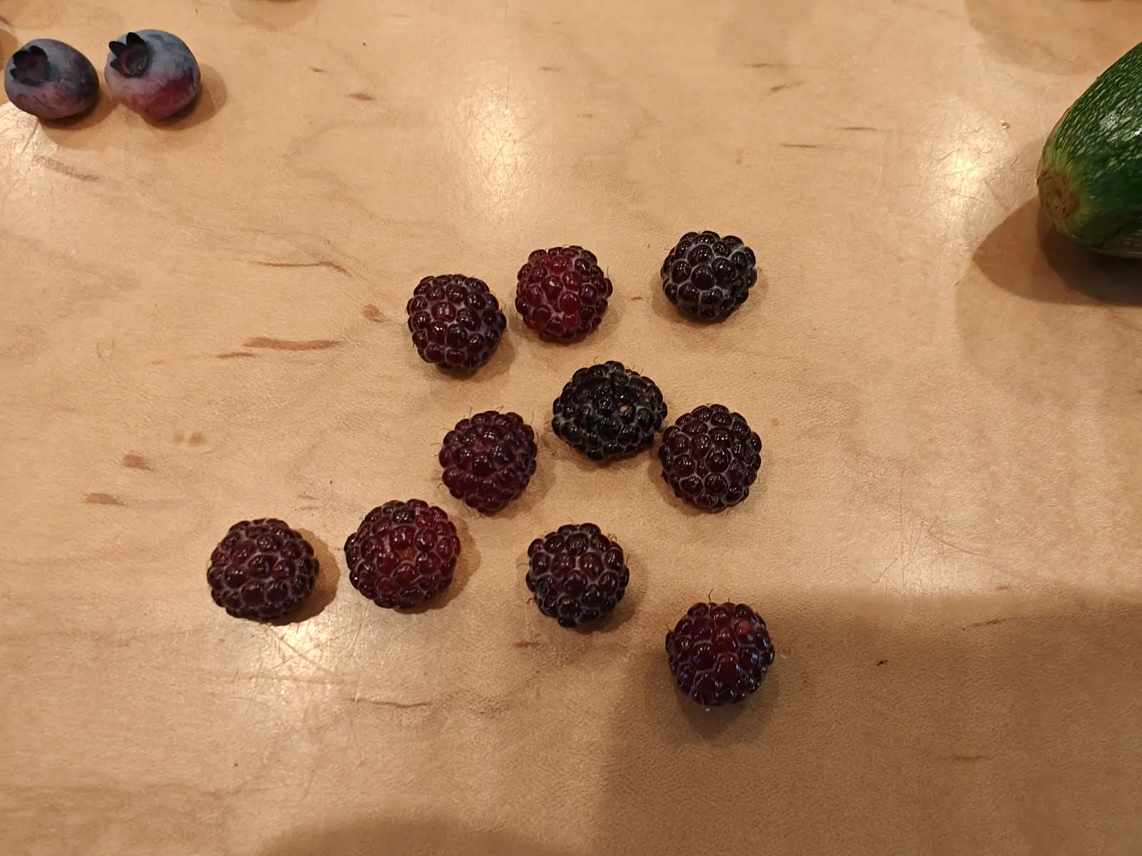 A scatter of dark red wineberries laid out on a pale wooden board with two blueberries in the top corner and the end of a zucchini entering the frame.