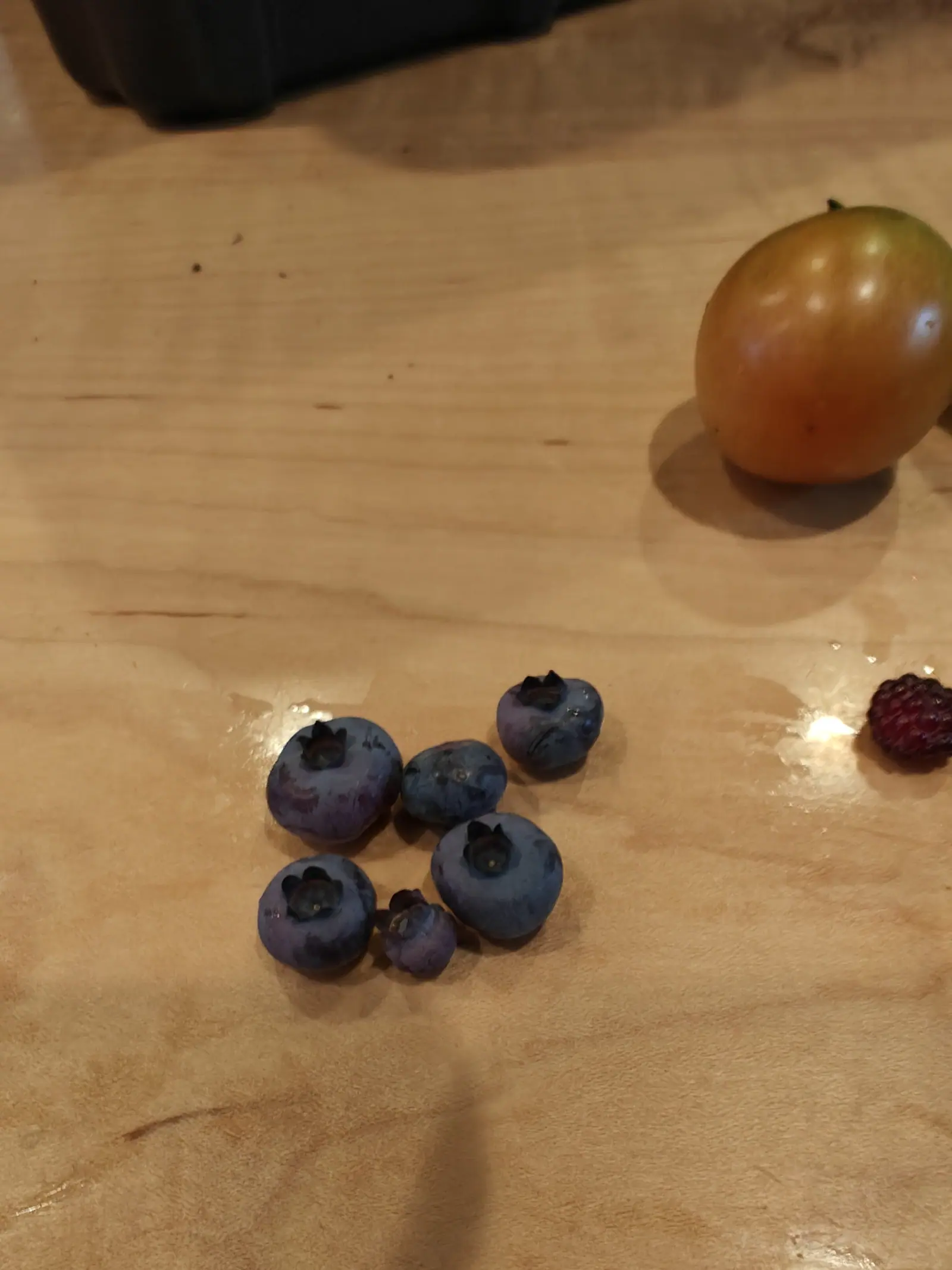 A handful of blueberries clustered on a pale wooden board with a yellow-green tomato above them and a single red berry at the right edge.