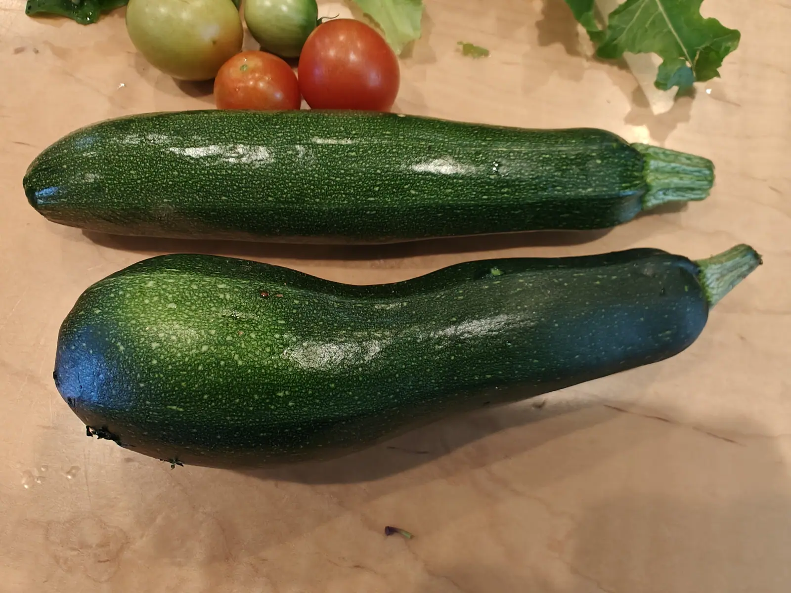 Two dark green zucchini lying side by side on a wooden board, one straight and one curved, with ripening red and green tomatoes behind.