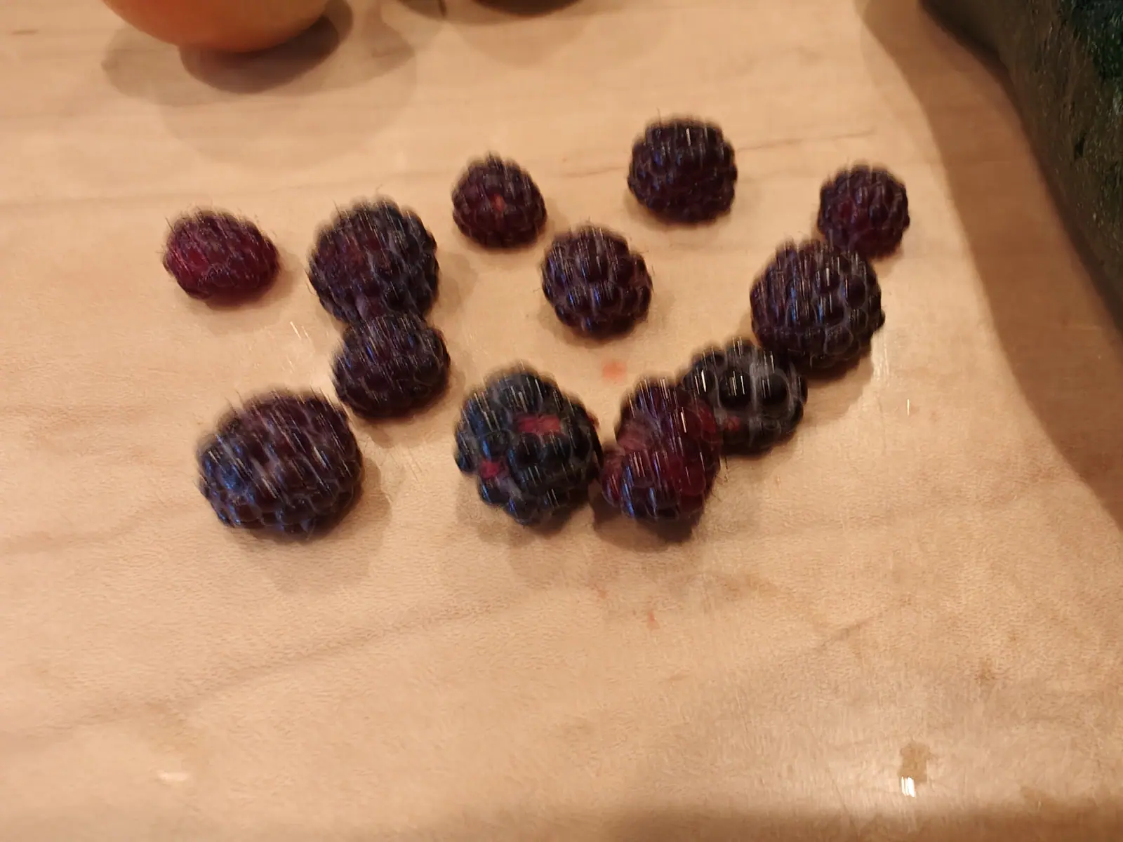 Wineberries scattered across a pale wooden board, a dozen small crimson-black fruits with tightly packed drupelets, a blurred orange fruit and a dark shape at the top edge.
