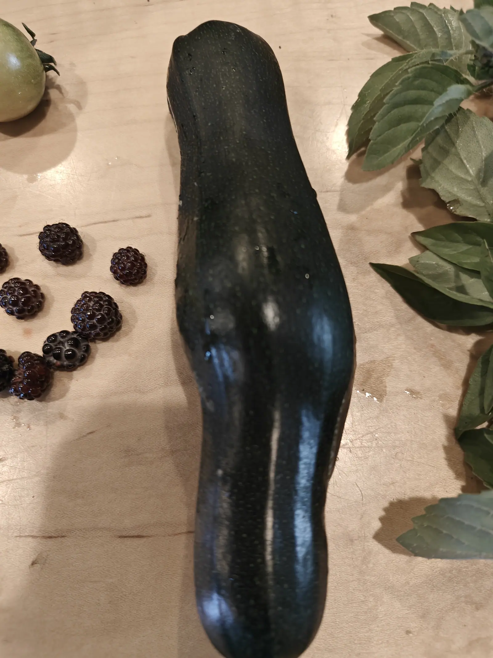 A single dark green zucchini lying on a pale wooden board, glossy and curved, with a green tomato, a scatter of near-black wineberries and cut sprigs of basil around it.