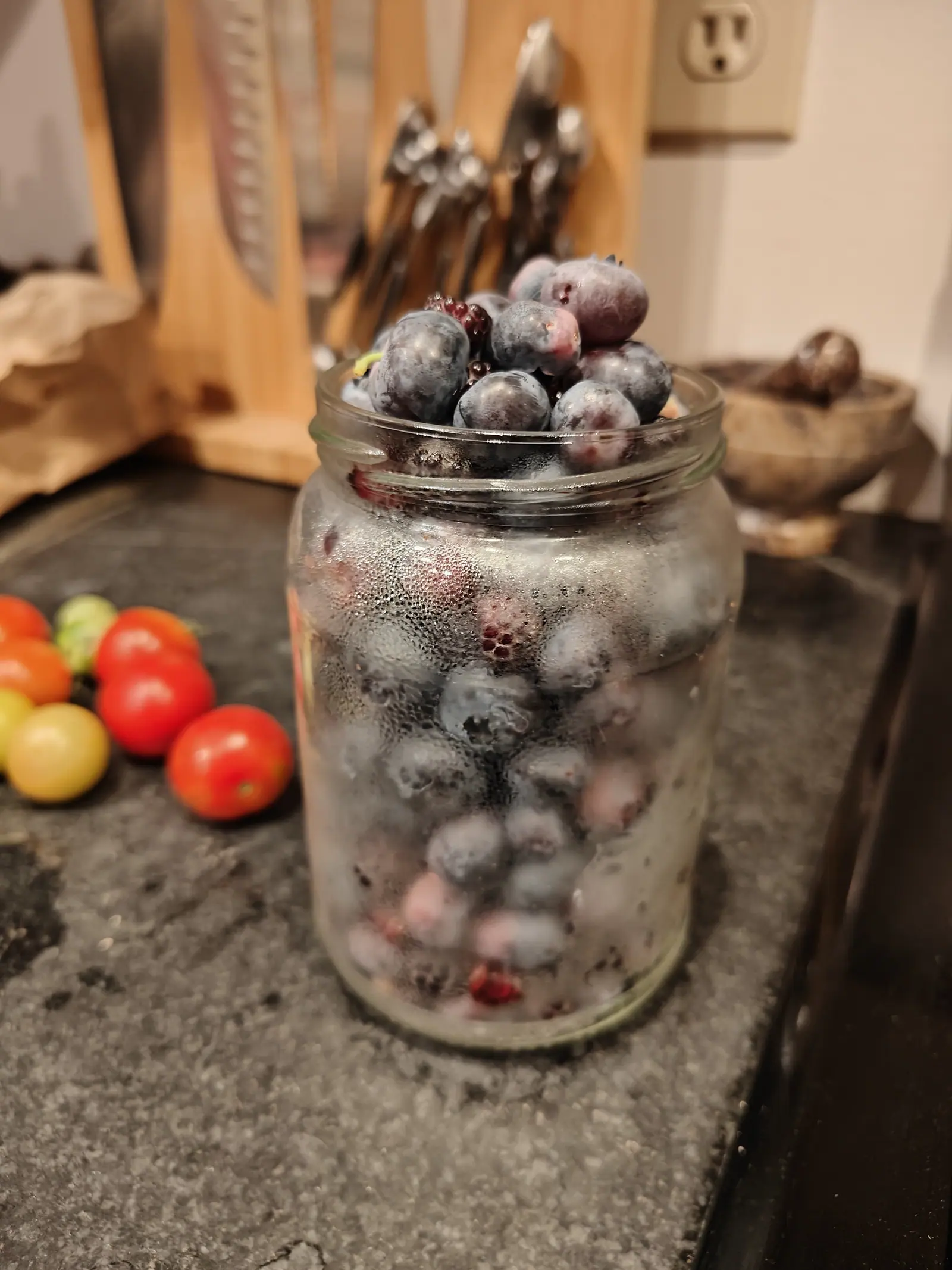 A glass jar packed with blueberries and a few dark wineberries on a dark kitchen counter, condensation on the glass, a knife block behind and small red and green tomatoes beside it.