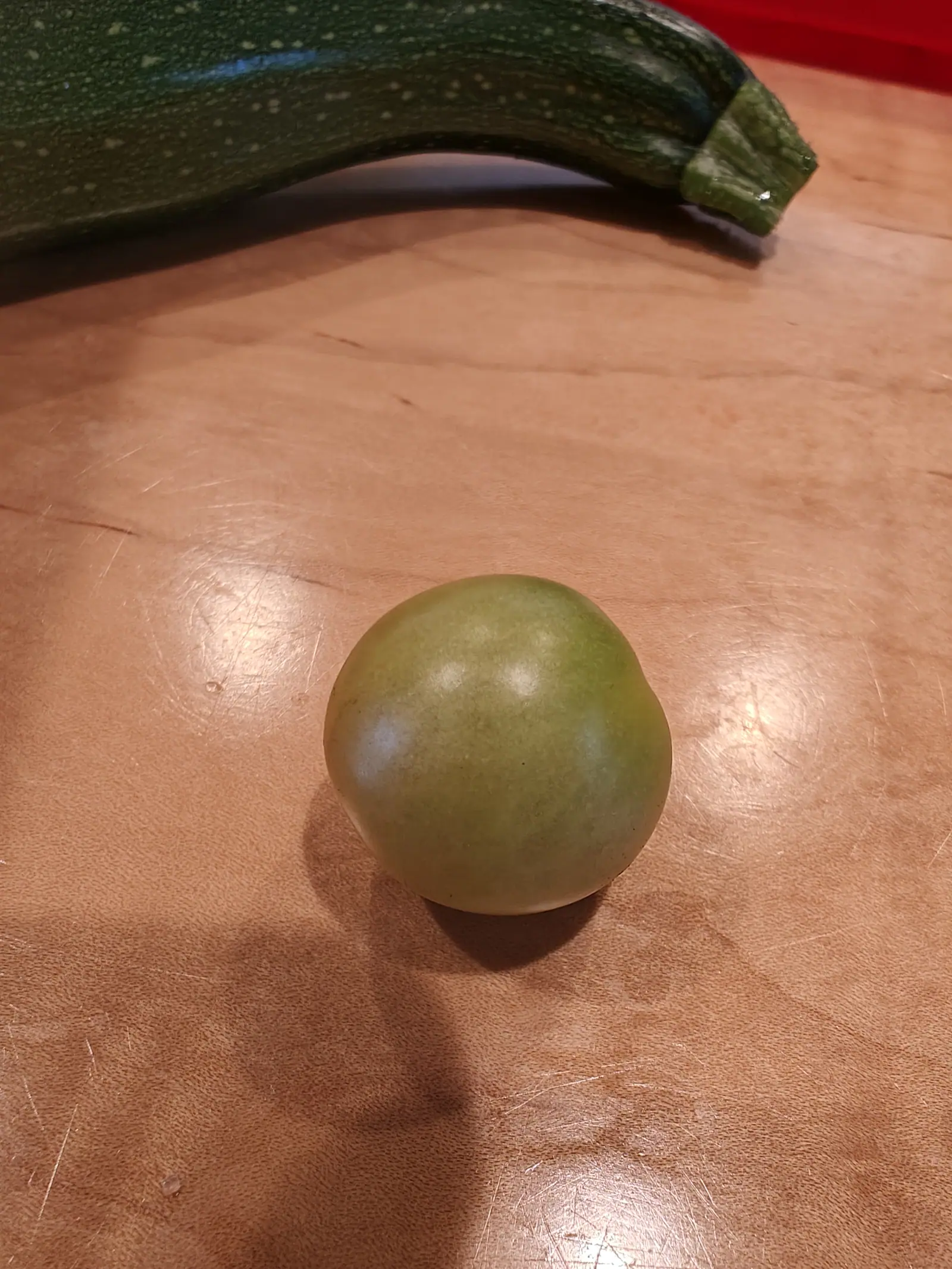 A single round green tomato on a scratched wooden chopping board under warm indoor light, a long dark green zucchini lying behind it.