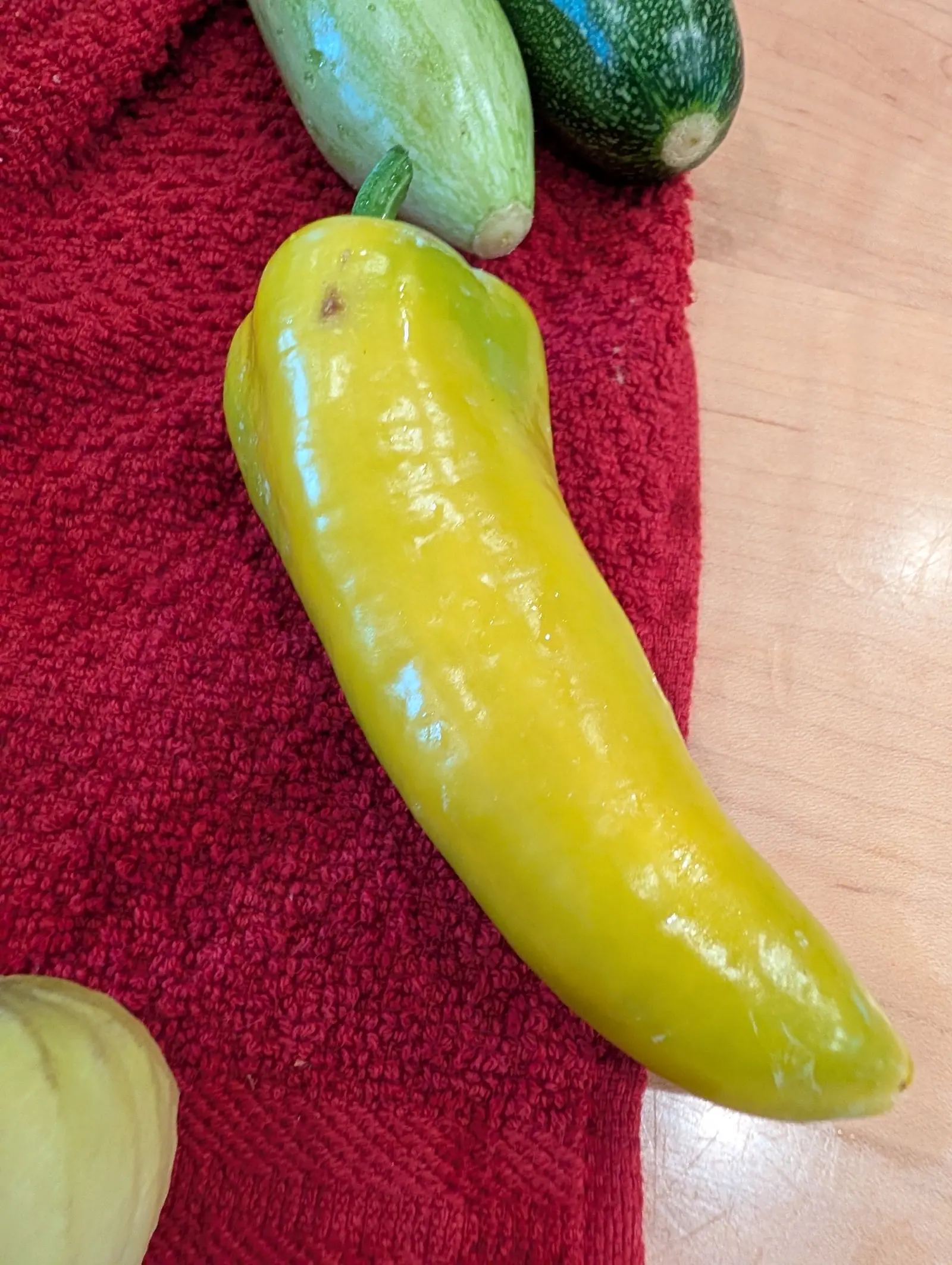 A large yellow pepper lying on a red towel on a wooden worktop, two small squash resting above it and a pale one at the corner of the frame.