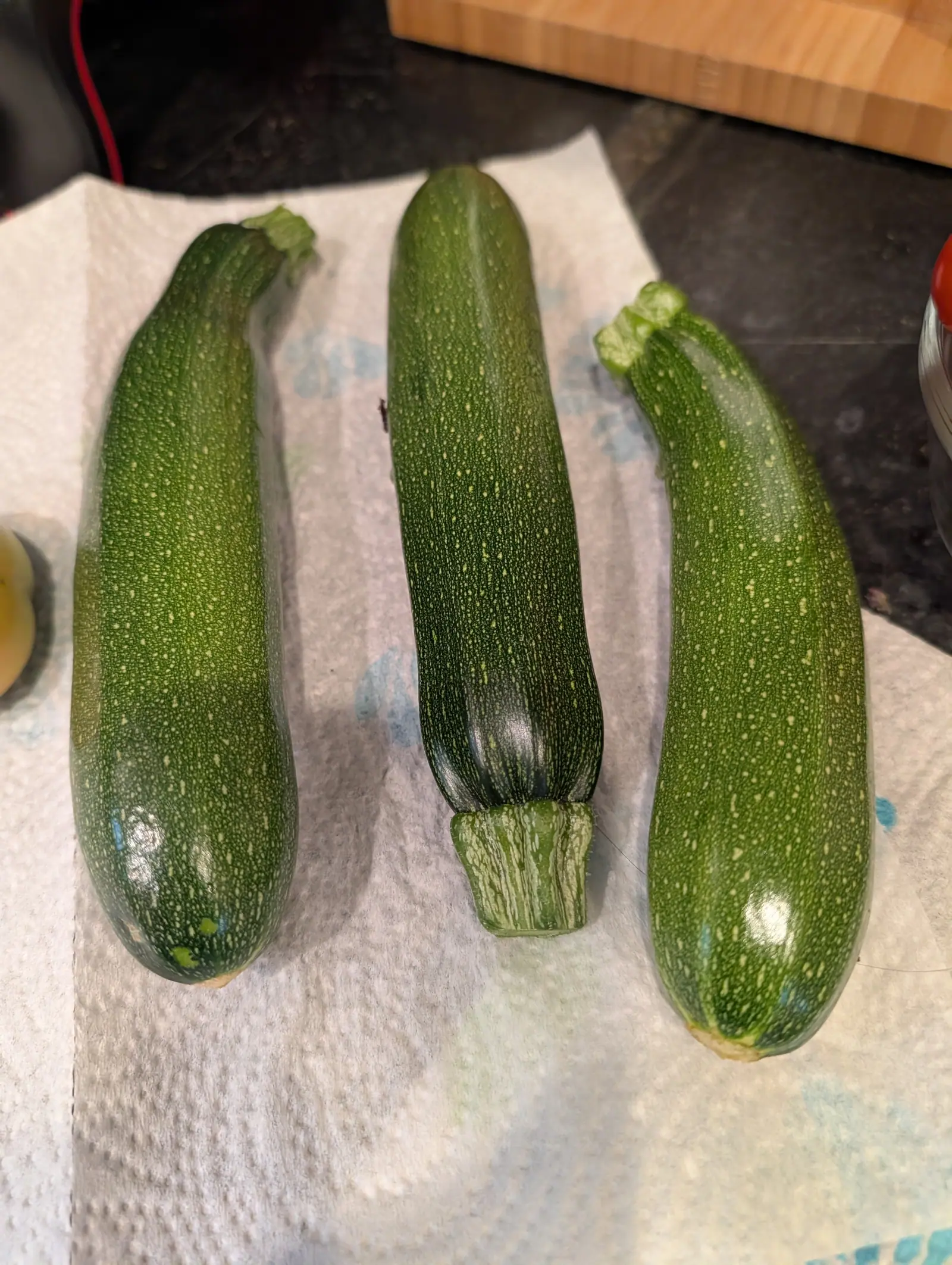 Three dark green zucchini laid side by side on kitchen paper on a dark worktop, their skins freckled pale, a wooden block behind.