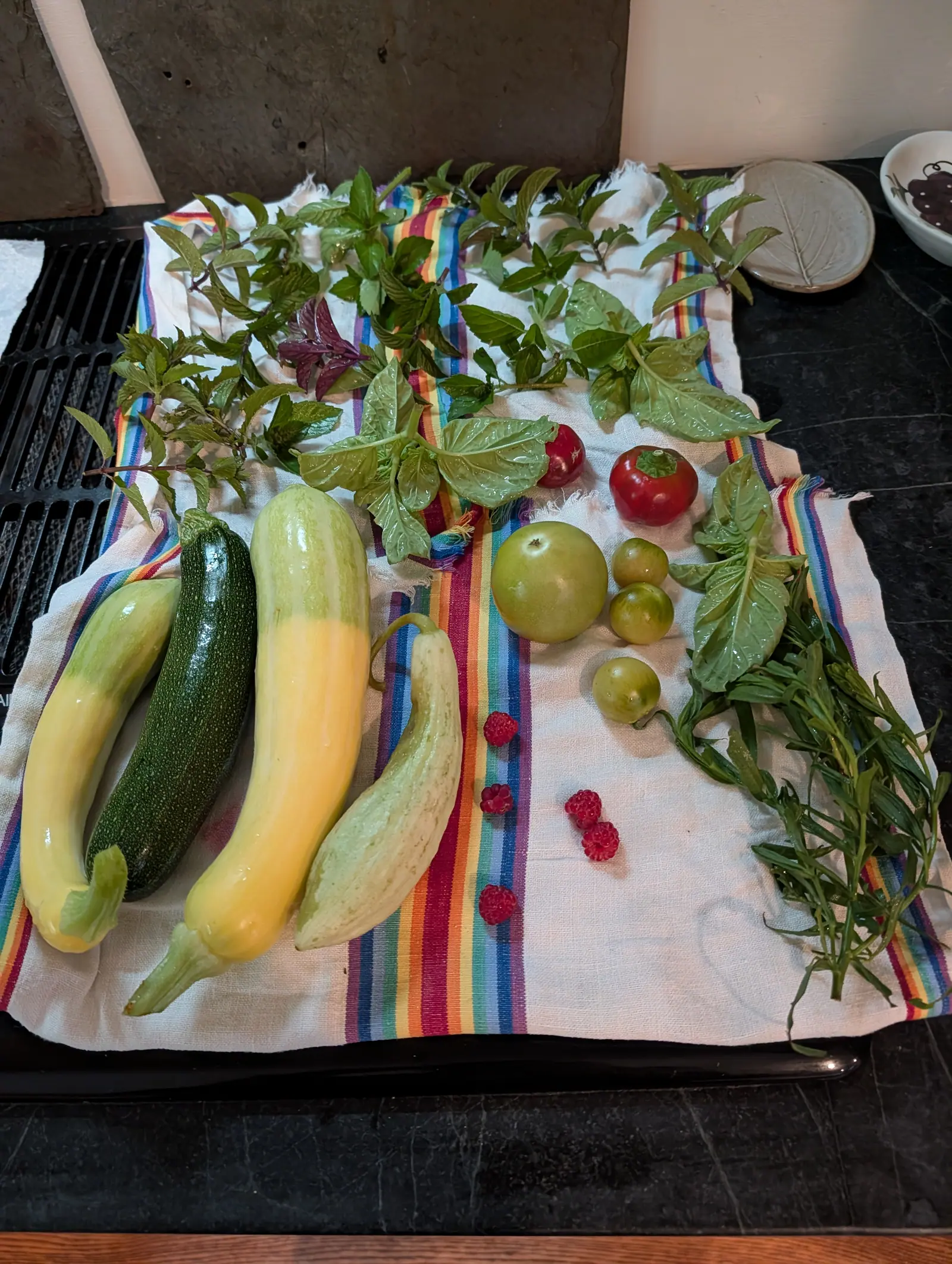 A mixed harvest laid out on a striped tea towel on a dark worktop: yellow and green zucchini, two red tomatoes and four green ones, small raspberries and bunches of basil, mint and tarragon.