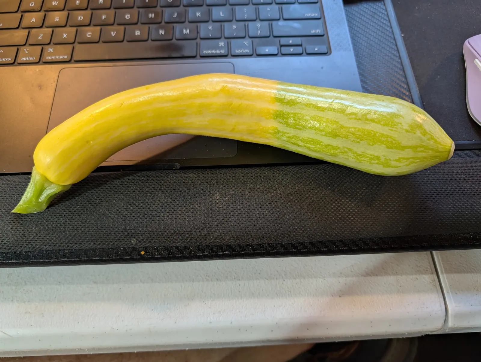 A long summer squash, yellow at the stem end and striped pale green at the tip, lying across the keyboard of an open laptop on a dark desk mat.