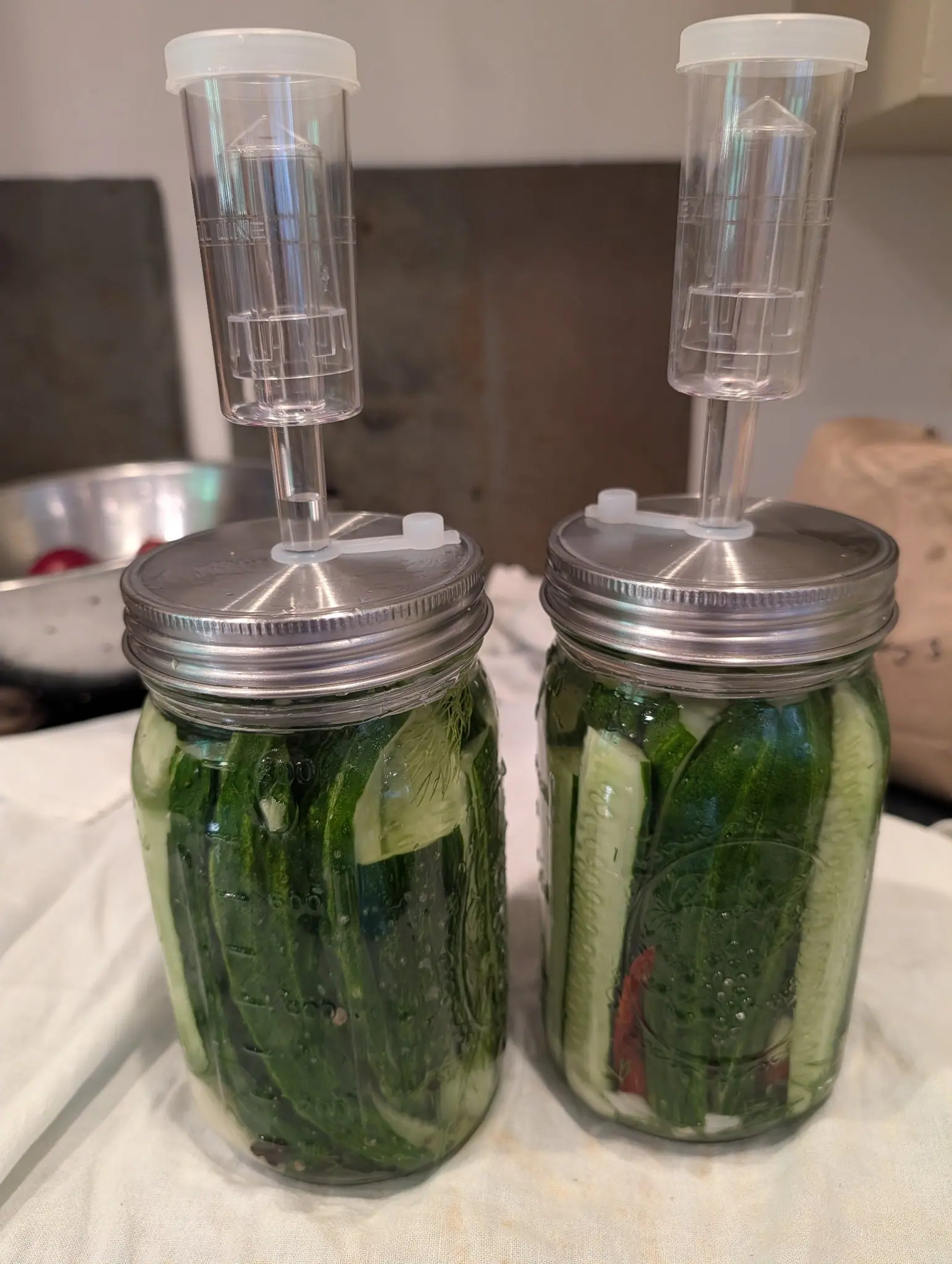 Two glass jars packed upright with whole cucumbers in brine, sealed with metal lids and tall clear plastic airlocks. Pale slices of onion and a red chilli show through the glass among the dark green skins. They stand on a white cloth, a metal bowl behind them.