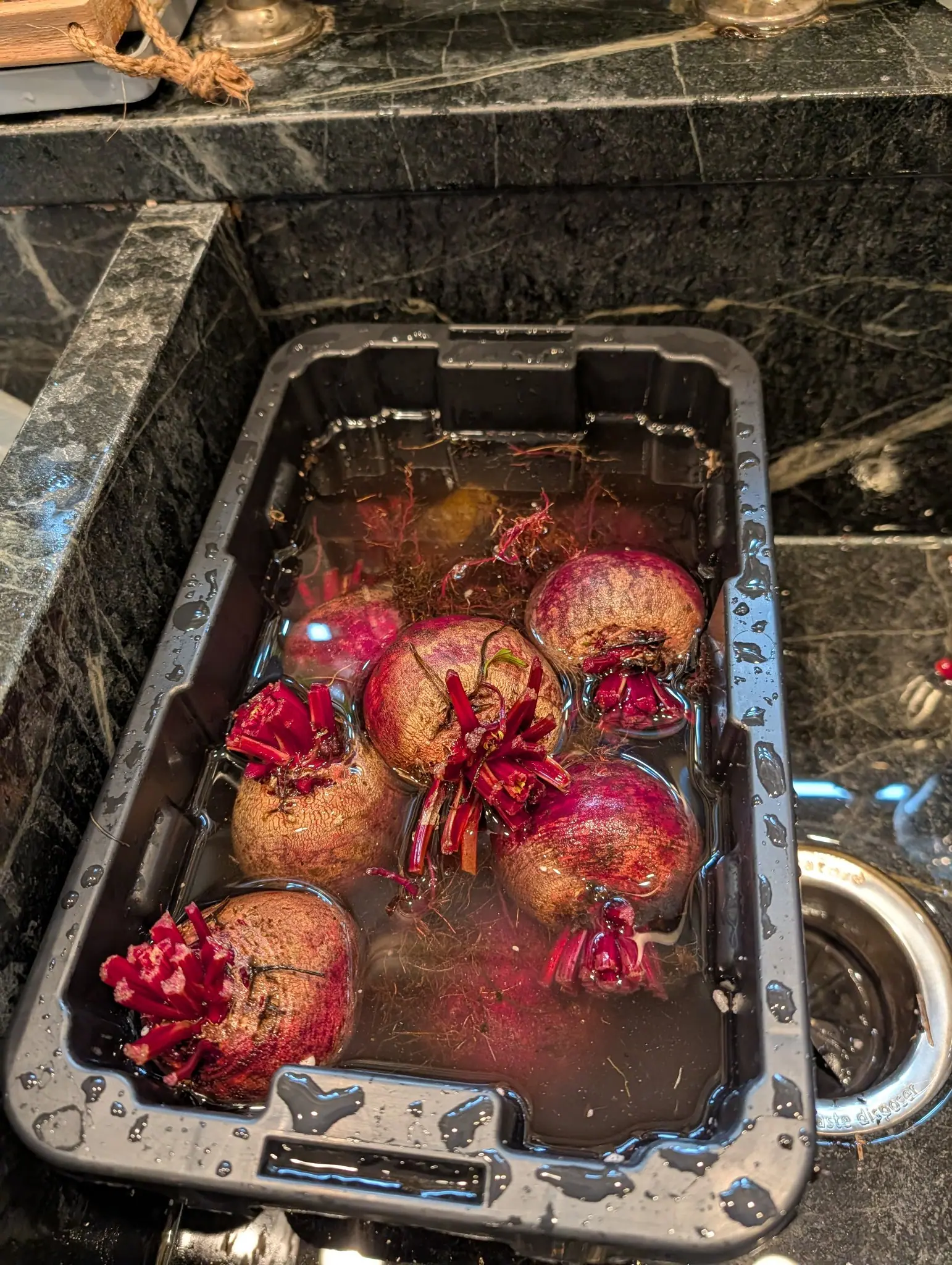 Beetroots soaking in a black plastic crate of water set into a dark stone sink, their trimmed magenta stalks and leaf bases floating around them.