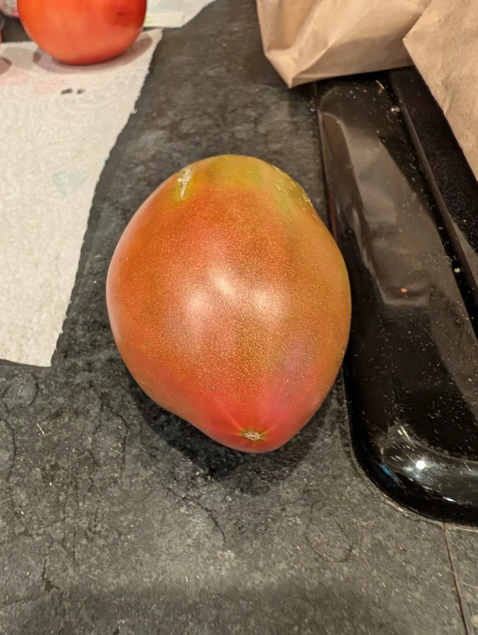 A large tomato ripening orange-red with green-purple shoulders, standing on a dark speckled worktop beside a black sink rim, a paper bag and a second tomato behind.