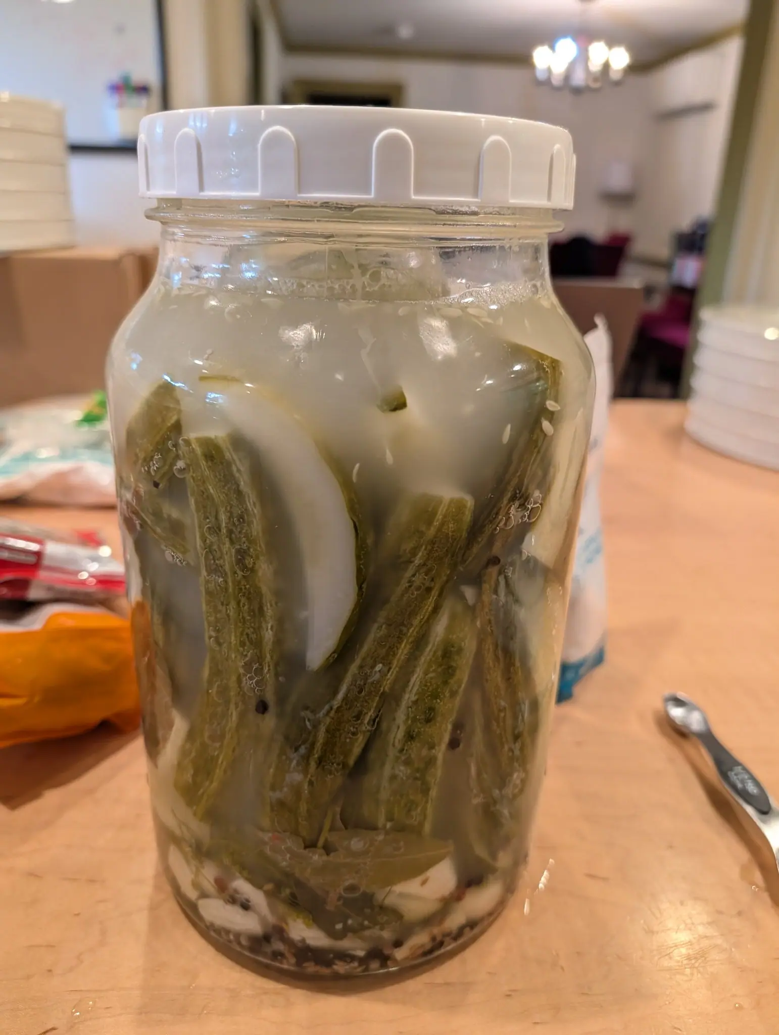 A glass preserving jar with a white lid standing on a wooden table, packed with ridged green pods, onion slices and garlic in cloudy brine, a dining room beyond.